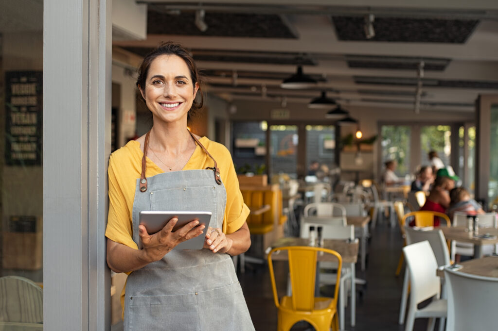 IT Support for business - Empowerment through technology. Portrait of happy woman standing at doorway of her store holding digital tablet. Cheerful mature waitress waiting for clients at coffee shop. Successful small business owner in casual clothing and grey apron standing at entrance and looking at camera.